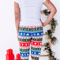 Back view of a woman in a white tee with a hand on the hip, wearing jogger pajama pants in red, green, blue, and white fair isle stripes with snowflakes, pine trees, and white dragon heads.