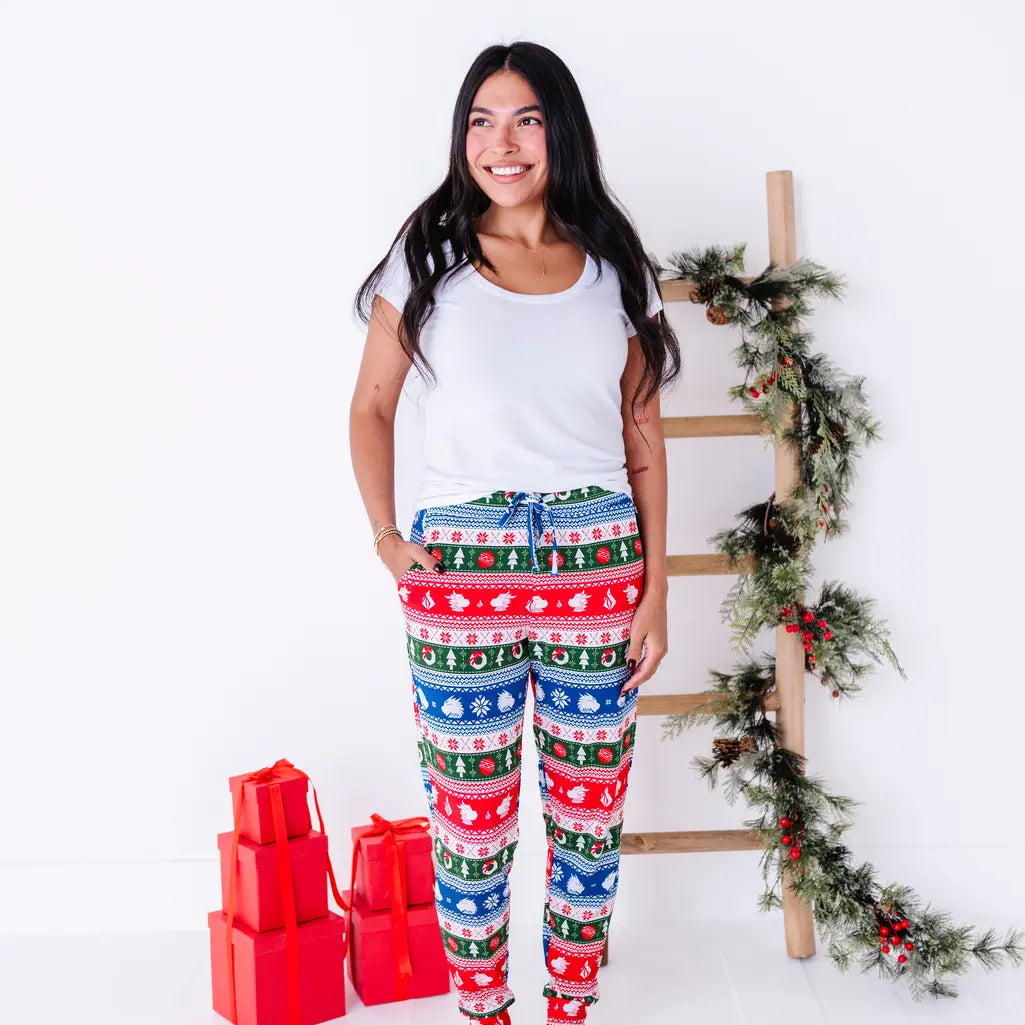 A woman smiling and looking to the side while standing in a white tee and jogger pajama pants in red, green, blue, and white fair isle stripes with snowflakes, pine trees, and white dragon heads.