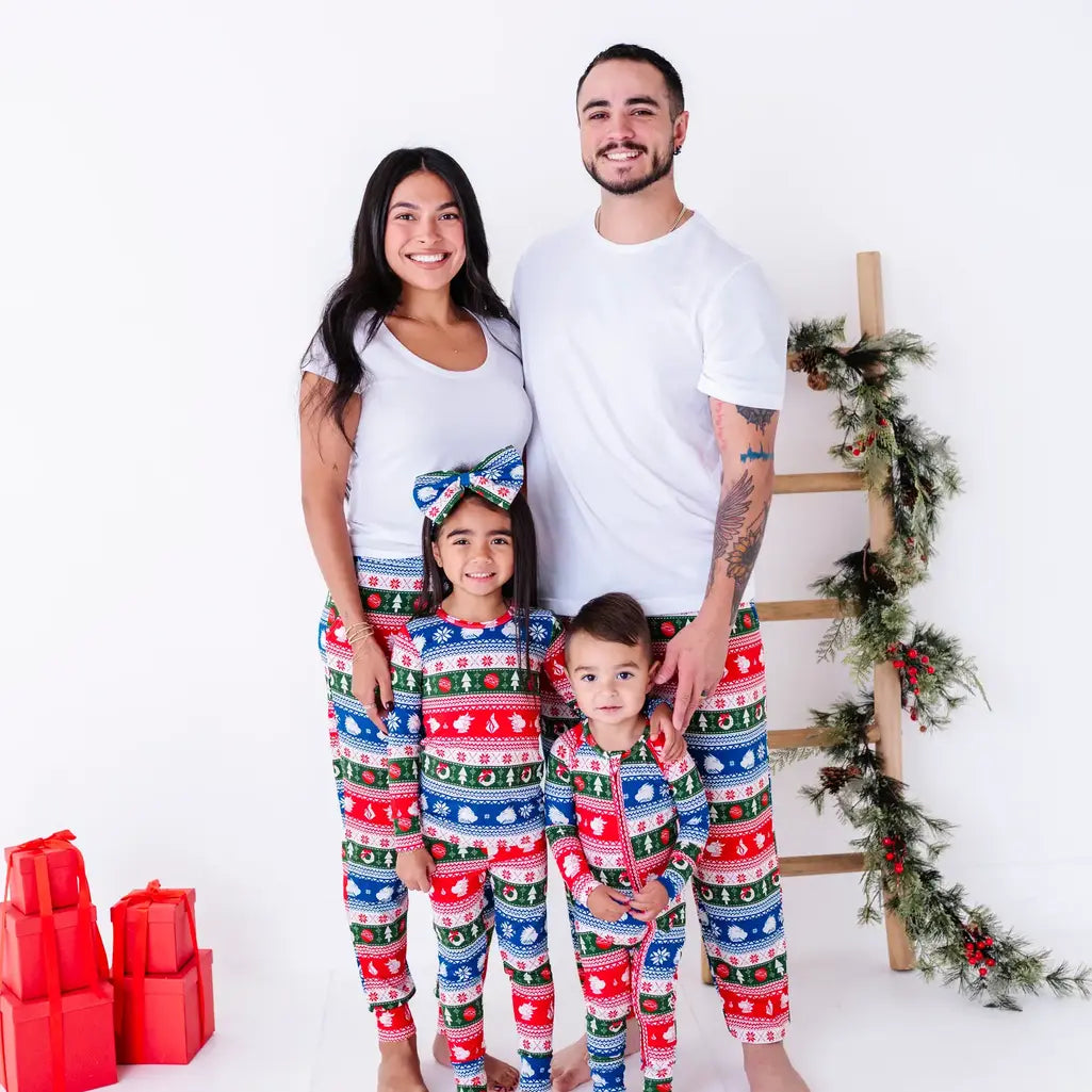 A family of four standing together and smiling at the camera in matching pajamas in red, green, blue, and white fair isle stripes with snowflakes, pine trees, and white dragon heads.
