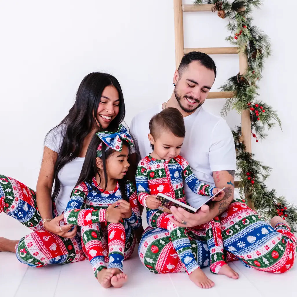 A family of four sitting on the floor reading a book together in matching pajamas in red, green, blue, and white fair isle stripes with snowflakes, pine trees, and white dragon heads.