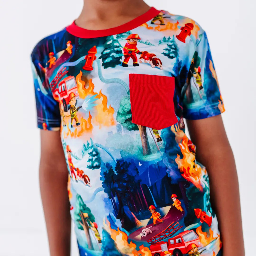 Close-up of a short-sleeve tee with a red chest pocket and a print of firefighters, fire trucks, and flames in a forest scene.
