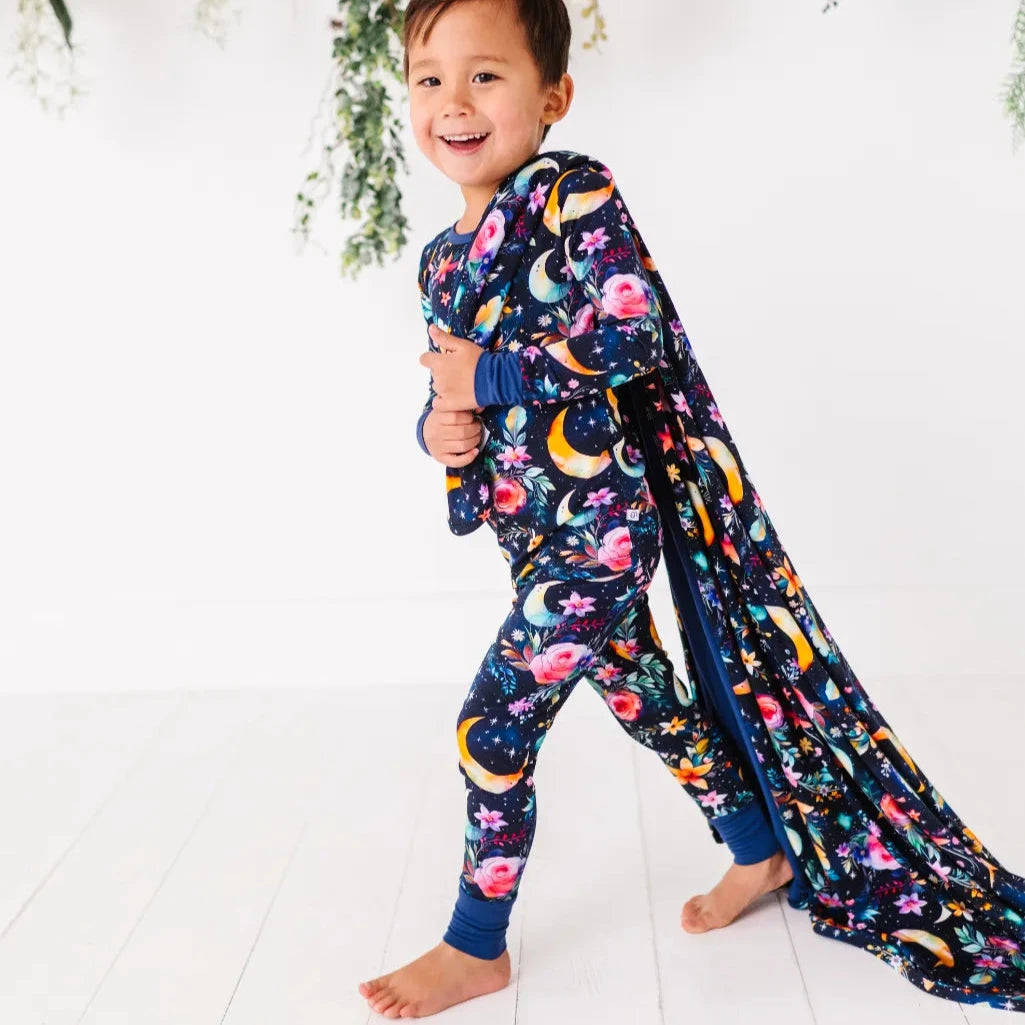 A child walking while pulling a matching blanket, wearing a navy pajama set with flowers, moons, and stars.
