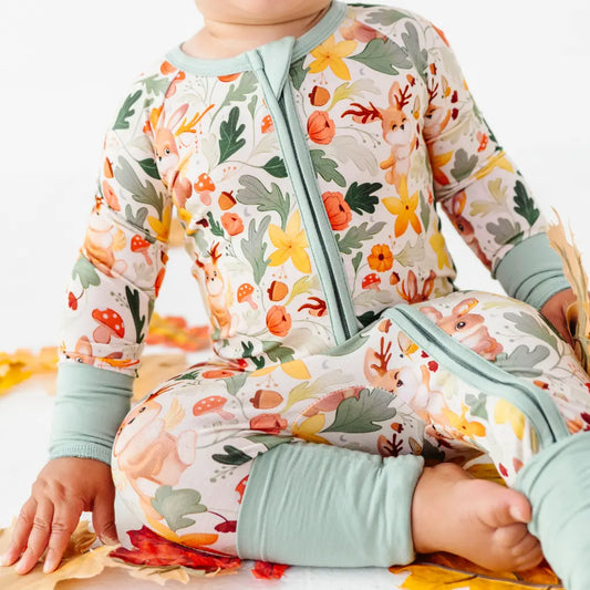 A cropped view of a baby seated on leaves in a zip up woodland print pajama with folded sage cuffs.