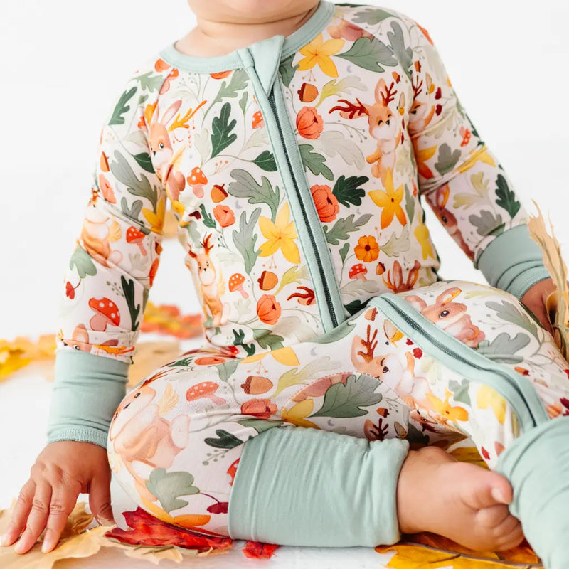 A cropped view of a baby seated on leaves in a zip up woodland print pajama with folded sage cuffs.