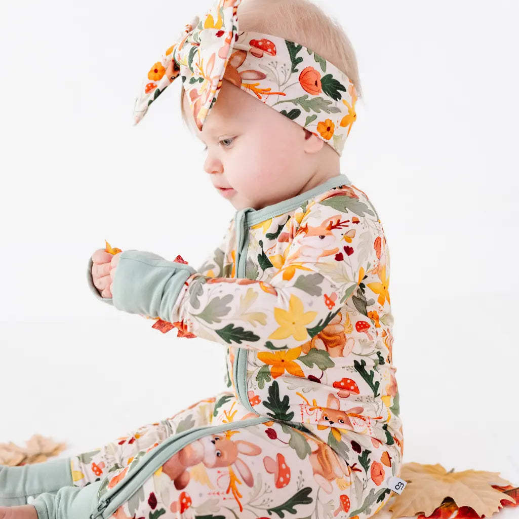 A side view of a baby in a zip up woodland print pajama and matching bow headband with sage cuffs.