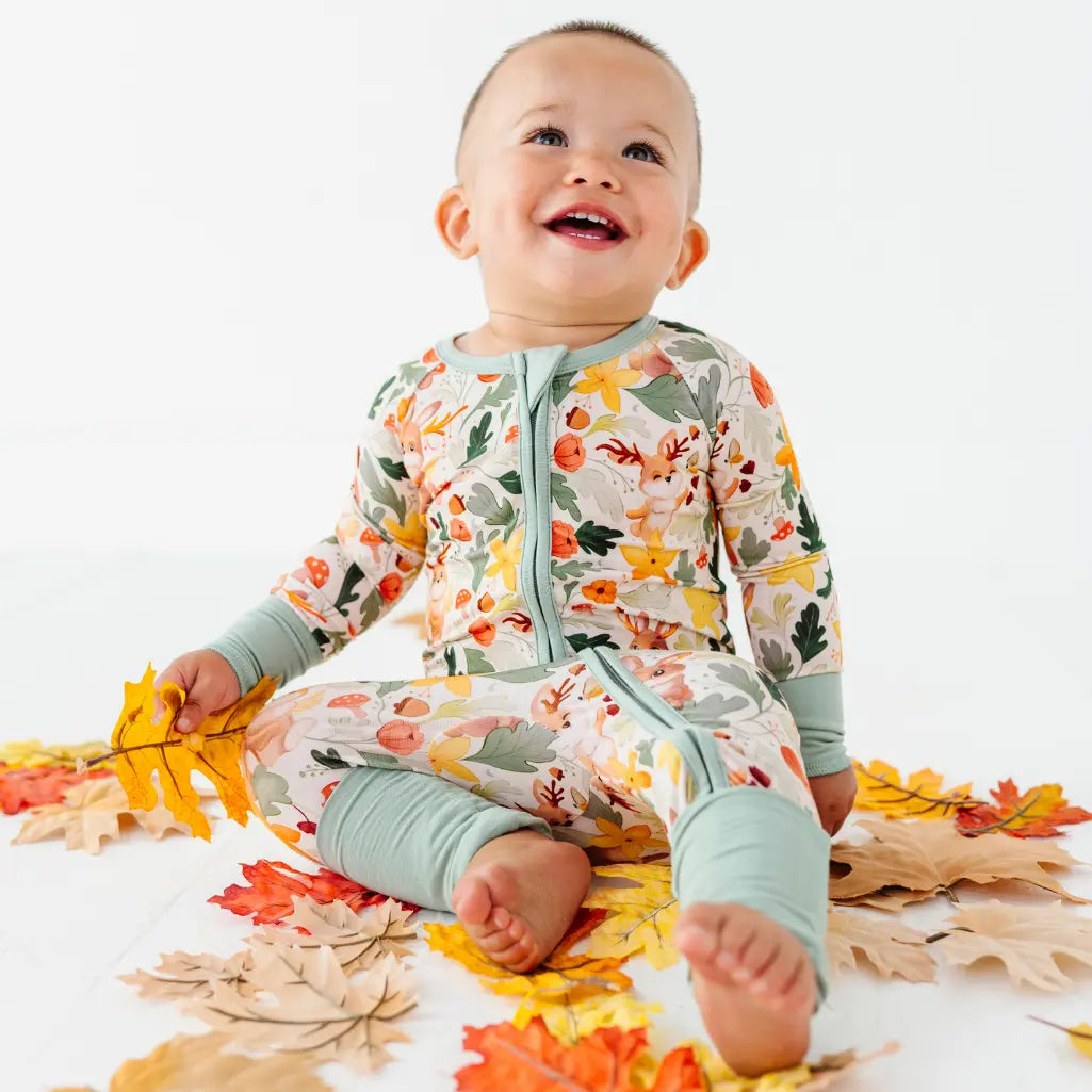 A baby sitting on autumn leaves holding one leaf while wearing a zip up woodland print pajama.
