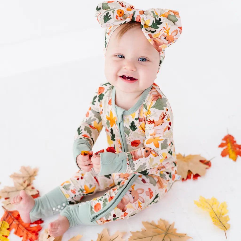 A baby sitting among autumn leaves in a zip up woodland print pajama with a matching bow headband.