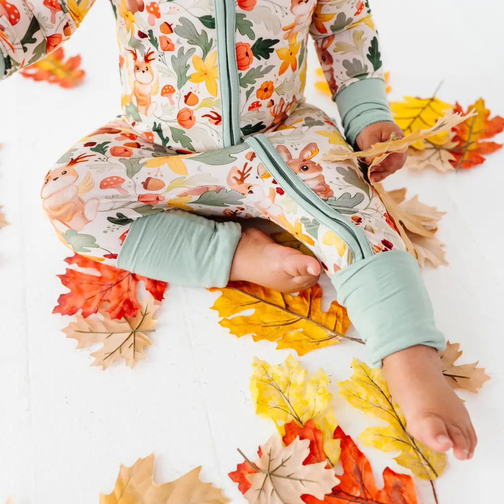 A close up of a baby seated on autumn leaves in a zip up woodland print pajama with fold over sage cuffs.