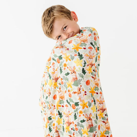 A child wrapped in a blanket with a woodland print of antlered bunnies, mushrooms, and yellow flowers on cream.