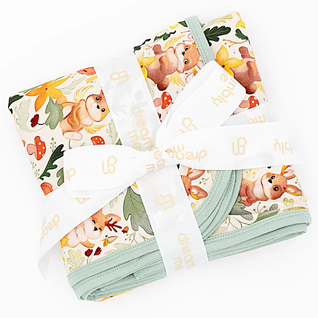 Two folded blankets with sage green trim tied with branded white ribbon on a white background in a woodland jackalope print.
