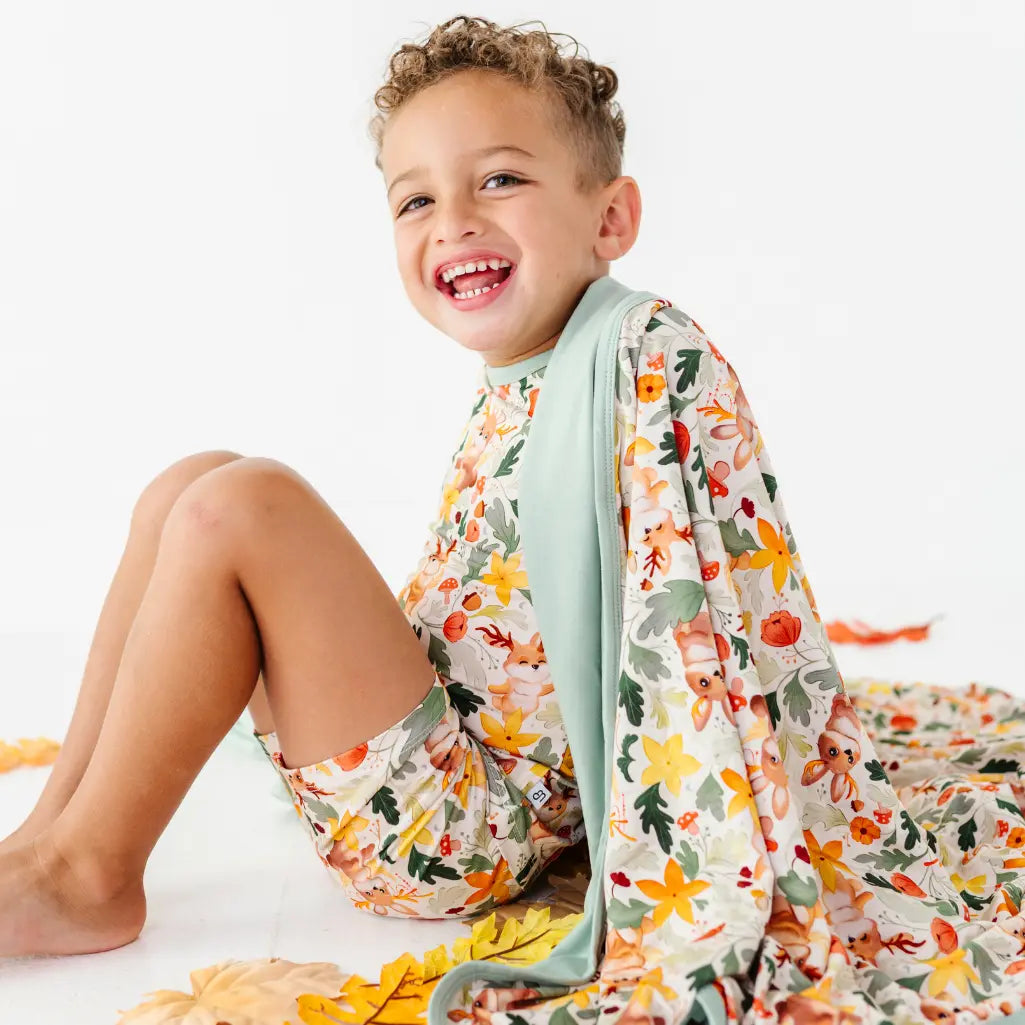 A child sitting among fall leaves with a woodland jackalope print blanket with sage green trim draped over one shoulder.