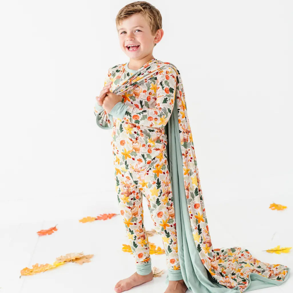 A child standing on a white floor with a woodland print blanket draped over one shoulder among fallen leaves.