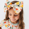 A close up of a child wearing a large woodland print bow headband with a matching pajama top.
