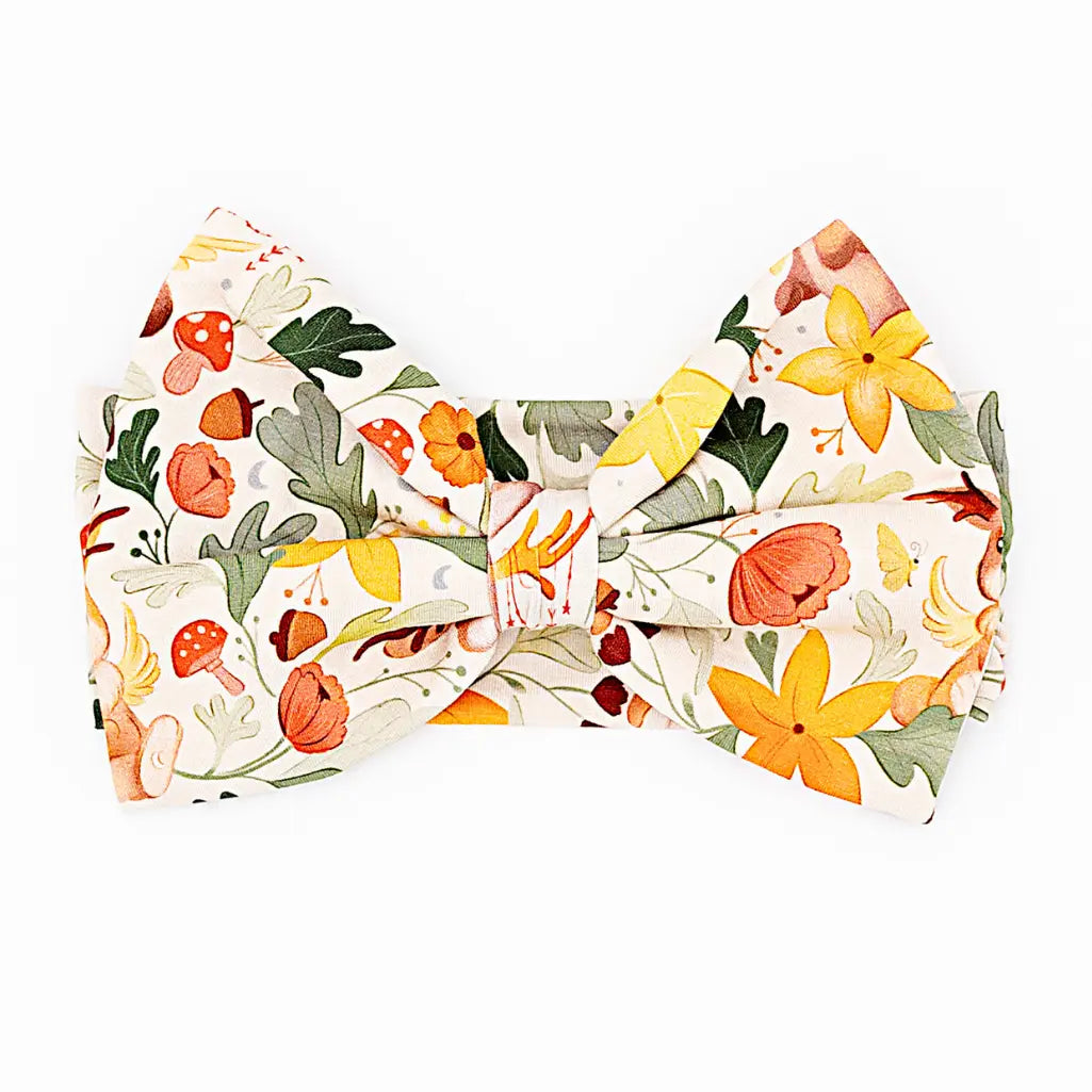 A large fabric bow headband laid flat on a white background in a cream woodland print with mushrooms, yellow flowers, and green leaves.