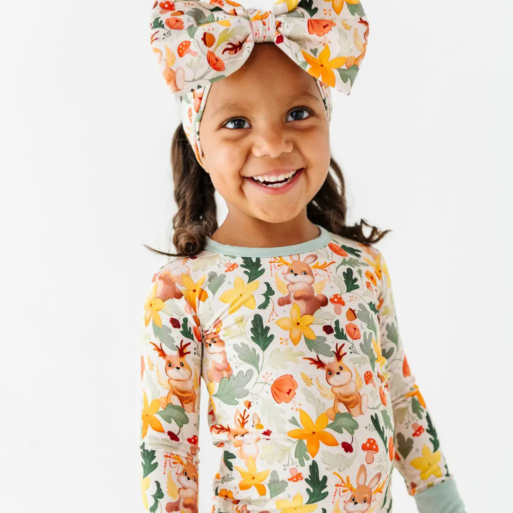 A child wearing a matching woodland print bow headband and long sleeve pajama top with sage neckline trim.