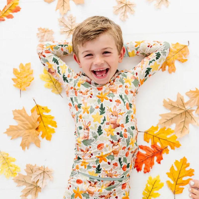A child lying on a white floor surrounded by autumn leaves in a woodland print pajama set with sage trim.