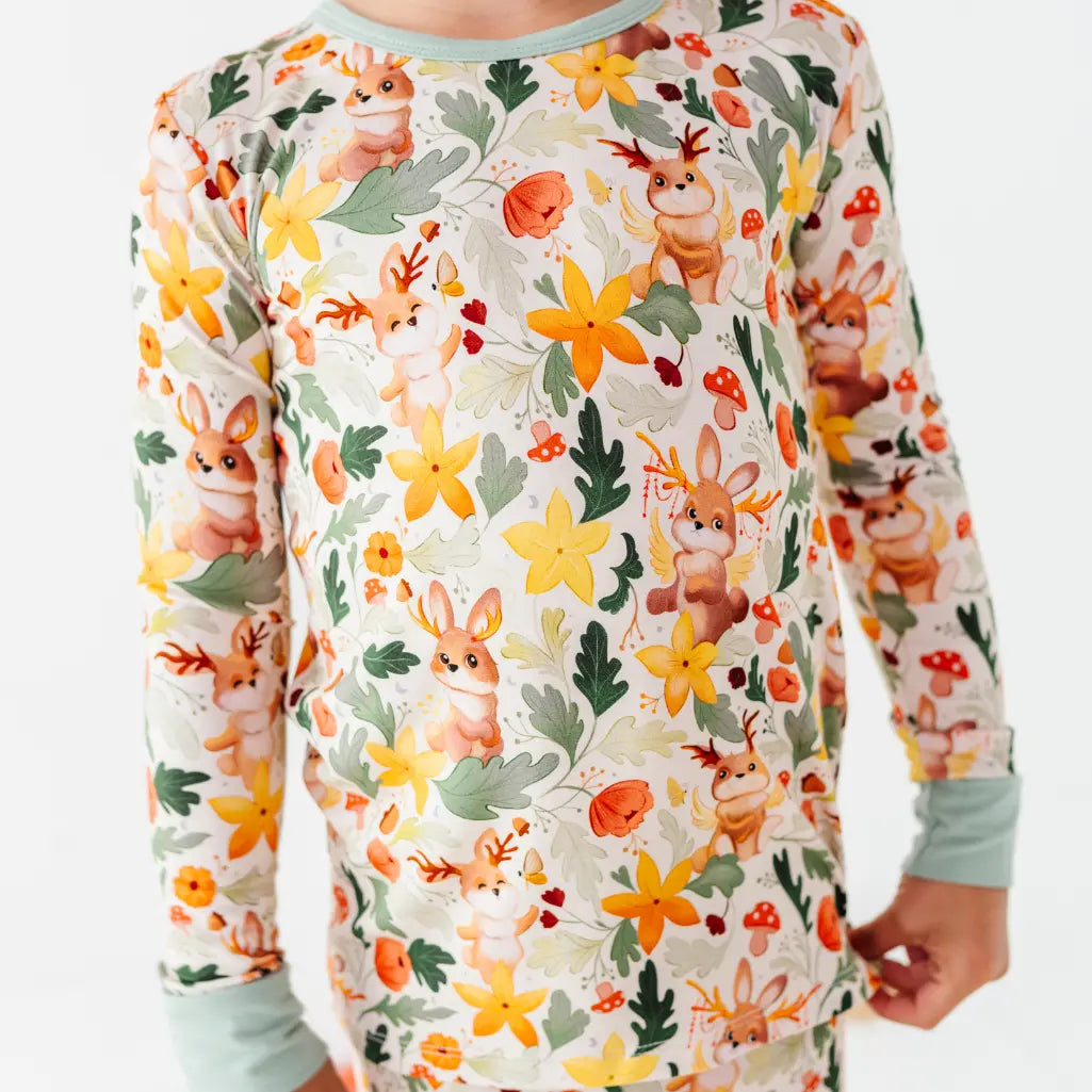 A close up of a pajama top showing antlered bunnies, mushrooms, and yellow flowers on cream with sage cuffs.