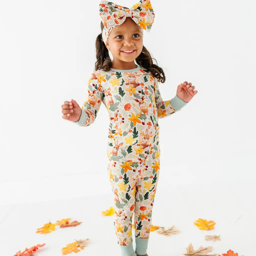 A child standing among fallen leaves in a woodland print one piece pajama with a matching bow headband.
