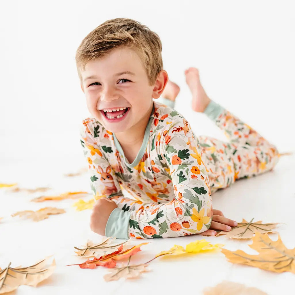 A child lying on their stomach among fallen leaves wearing a woodland print pajama set with sage cuffs.