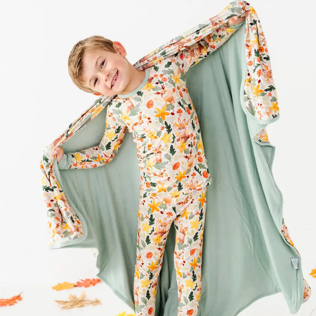 A child in a matching two piece pajama holding open a woodland print blanket with a sage green backing.