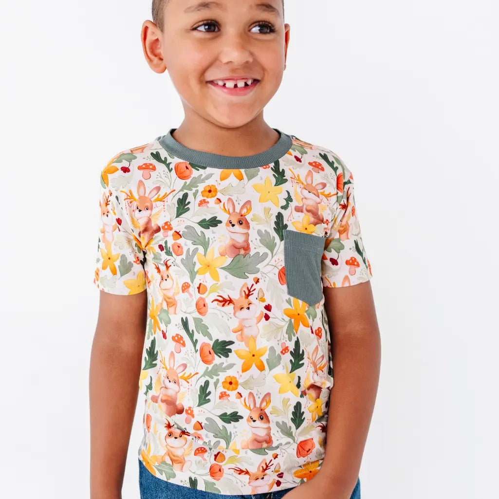 A smiling child on a white background wearing a short sleeve pocket tee with a dark green collar in a woodland jackalope print.