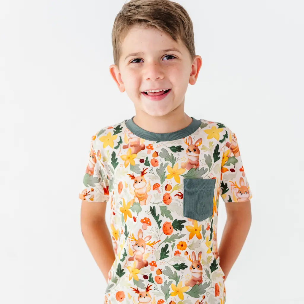A smiling child on a white background wearing a woodland jackalope print pocket tee with a dark green collar.