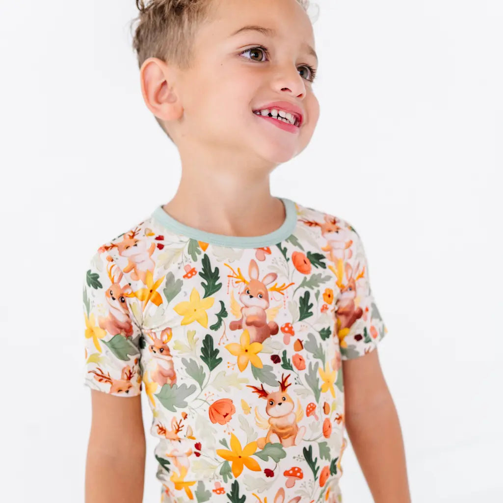 A smiling child looking to the side wearing a short sleeve pajama top with sage trim in a woodland jackalope print.