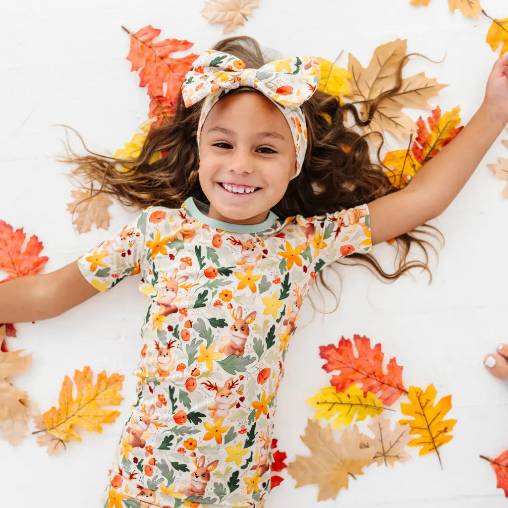 A child lying on a white floor surrounded by fall leaves wearing a woodland jackalope print pajama top and bow headband.