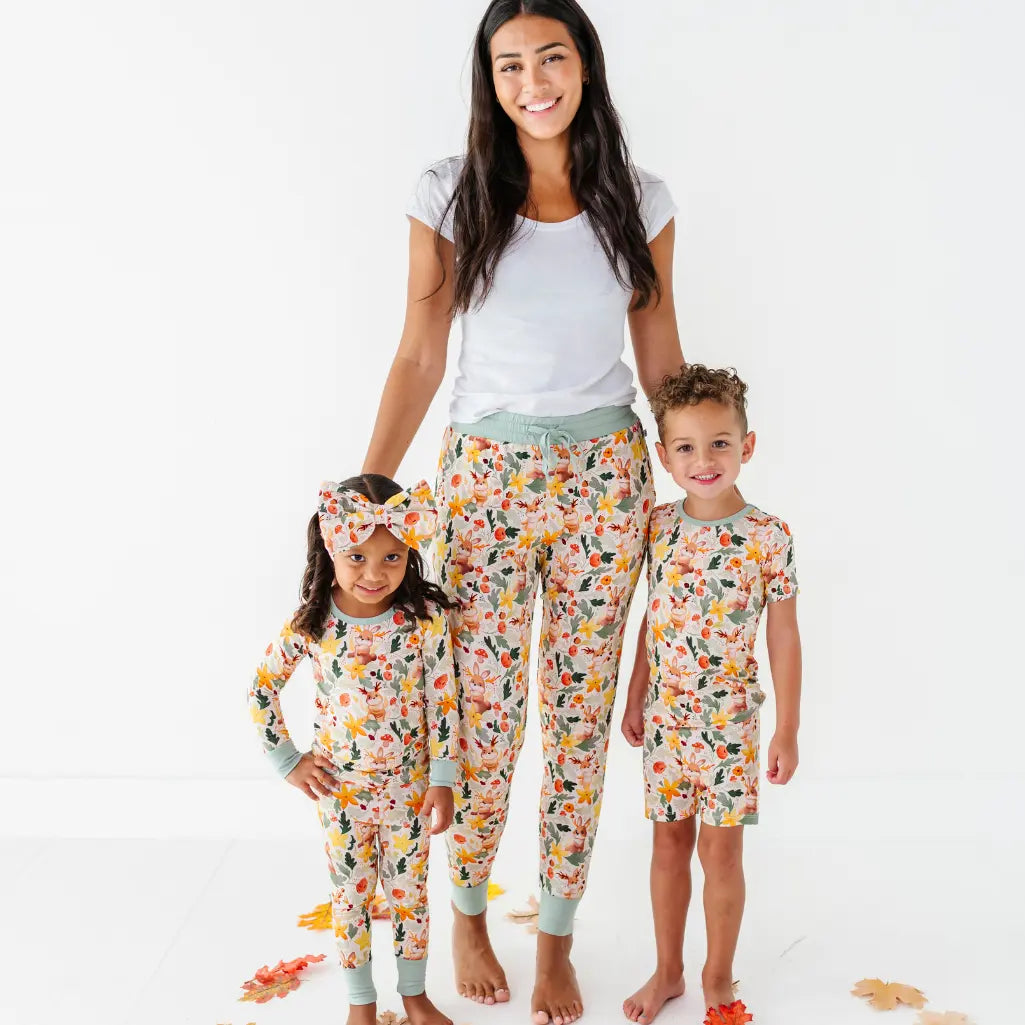An adult in woodland print pajama pants stands between two children in matching long sleeve and short pajama sets.
