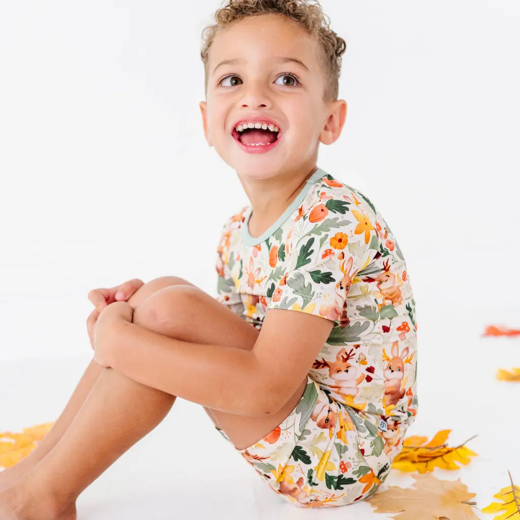 A child sitting on a white floor beside fall leaves wearing a woodland jackalope print short sleeve pajama set.