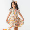 A child holding out the skirt of a woodland jackalope print twirl dress with a matching bow headband among fallen leaves.