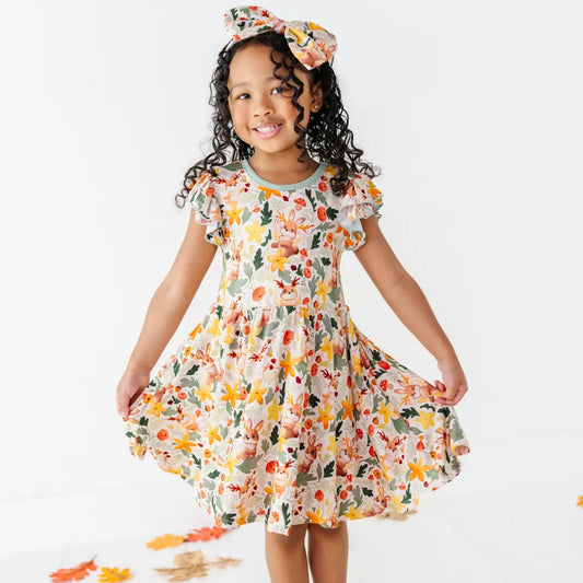 A child holding out the skirt of a woodland jackalope print twirl dress with a matching bow headband among fallen leaves.