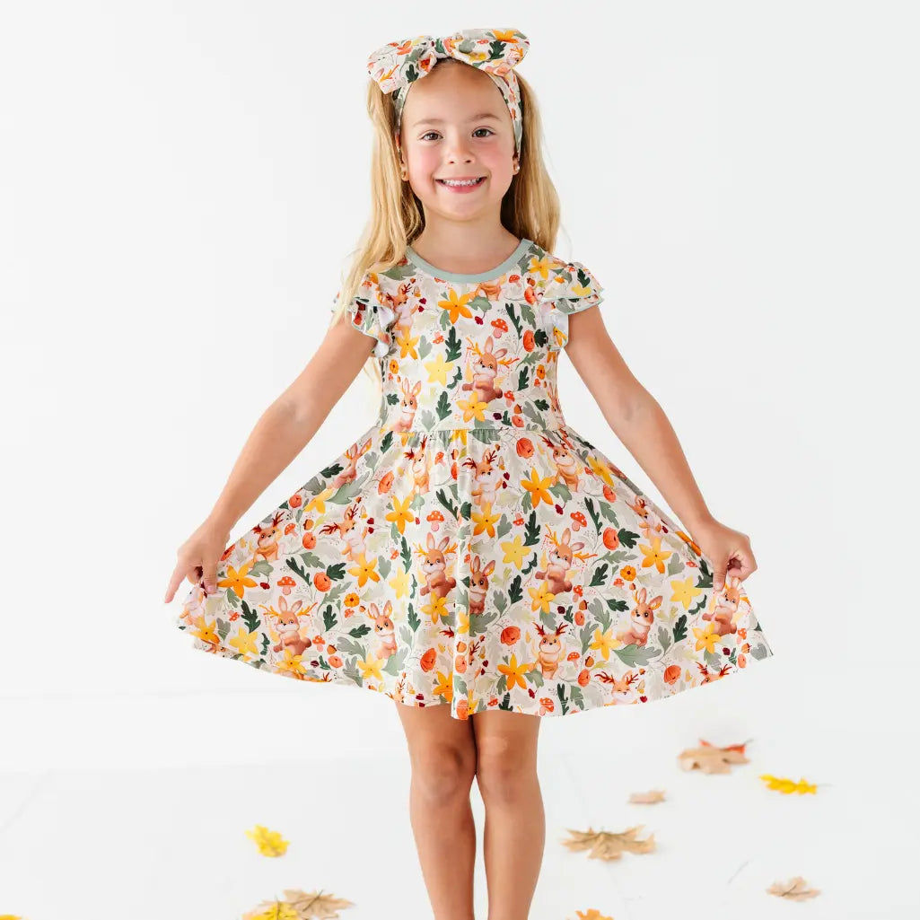A child holding out the skirt of a woodland jackalope print twirl dress with a matching bow headband among fallen leaves.