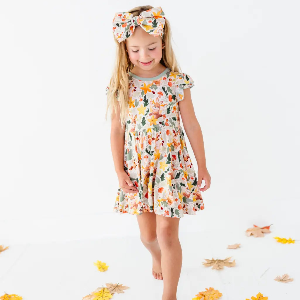A child walking barefoot among fallen leaves in a woodland jackalope print twirl dress with a matching bow headband.