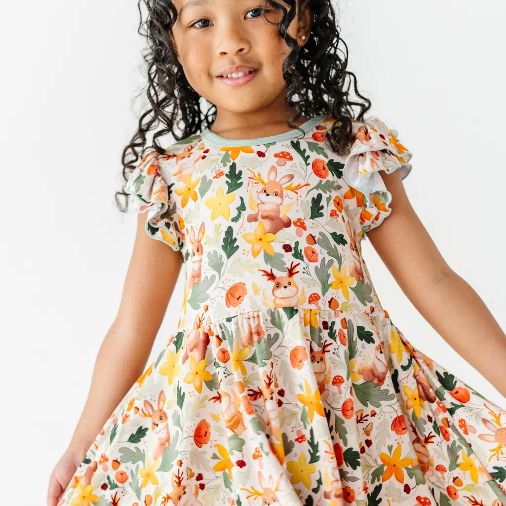 A child wearing a flutter sleeve twirl dress with sage trim in a woodland print with antlered bunnies and yellow flowers.