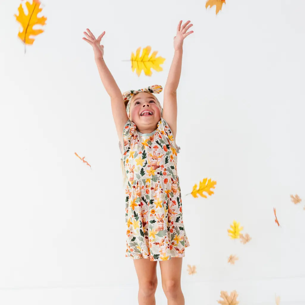 A child with arms raised tossing fall leaves while wearing a woodland jackalope print twirl dress and matching bow headband.