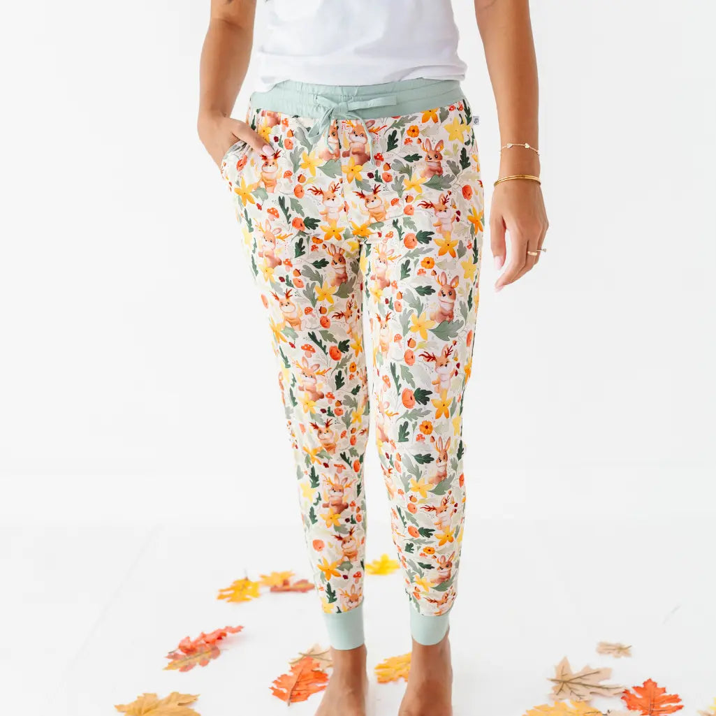An adult standing among fallen leaves with hands in the pockets of woodland print pajama pants.