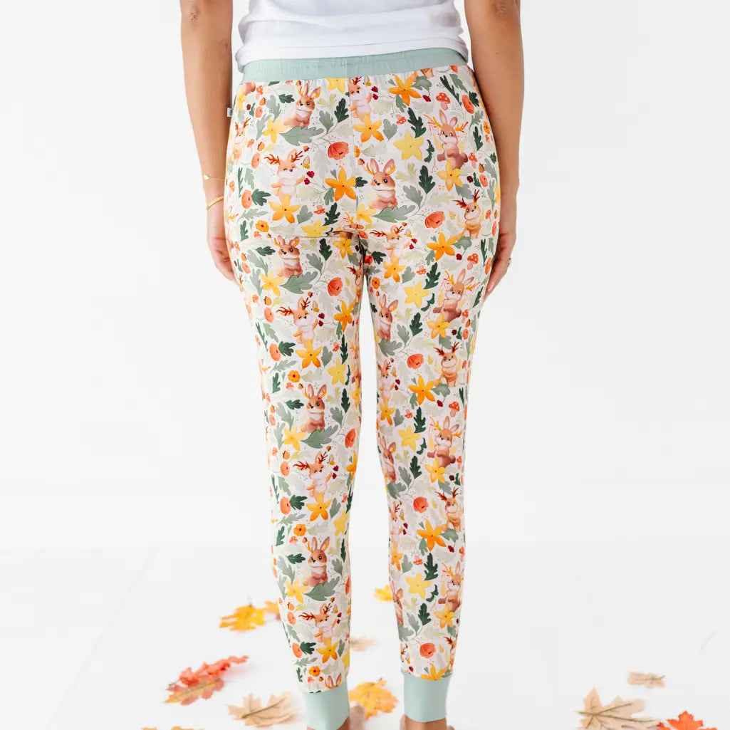 A back view of an adult wearing woodland print pajama pants with sage ankle cuffs among fallen leaves.