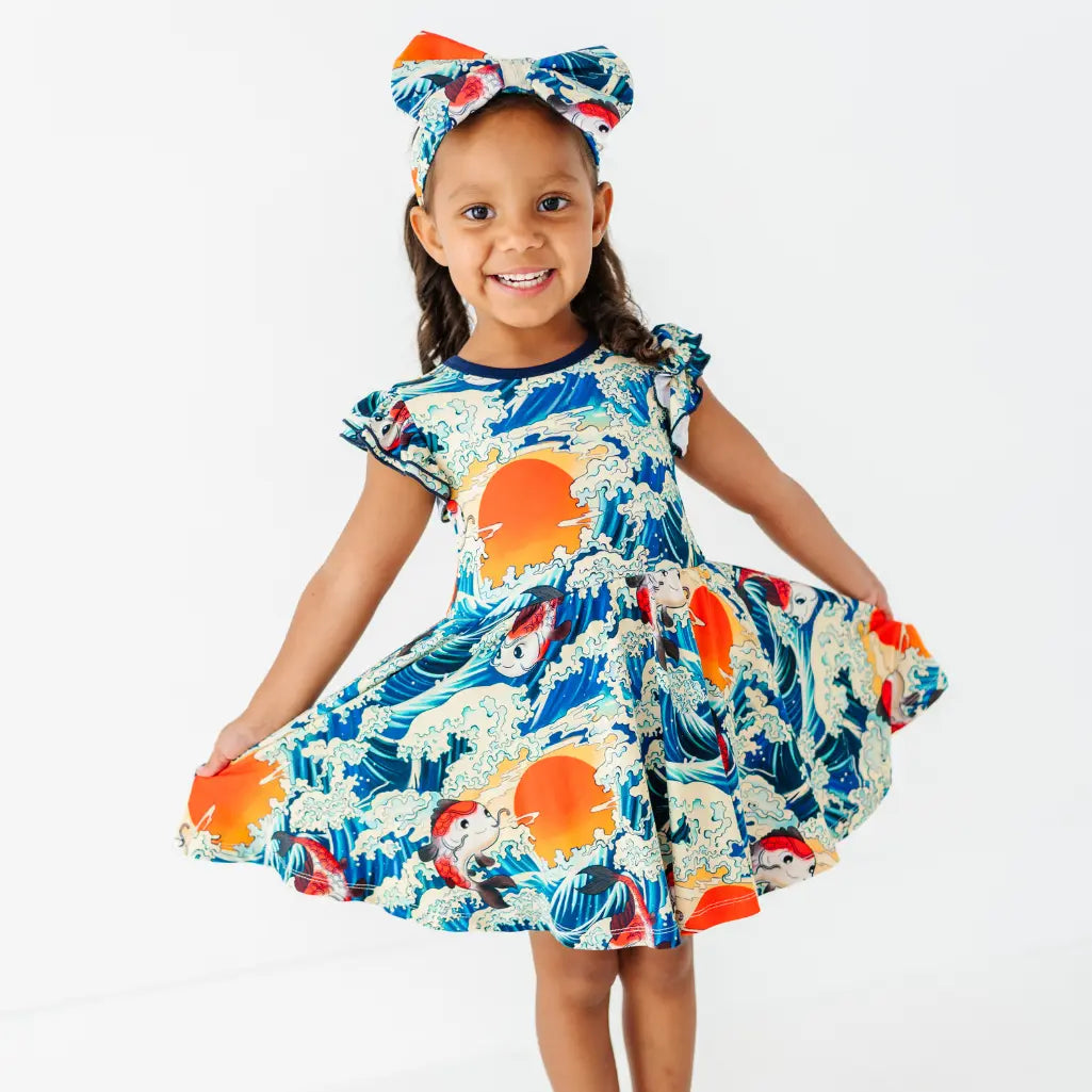 A smiling child in a matching bow headband holding out the skirt of a flutter sleeve twirl dress with a koi fish, wave, and sunset print.