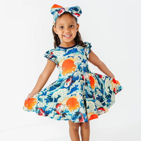 A smiling child in a matching bow headband holding out the skirt of a flutter sleeve twirl dress with a koi fish, wave, and sunset print.