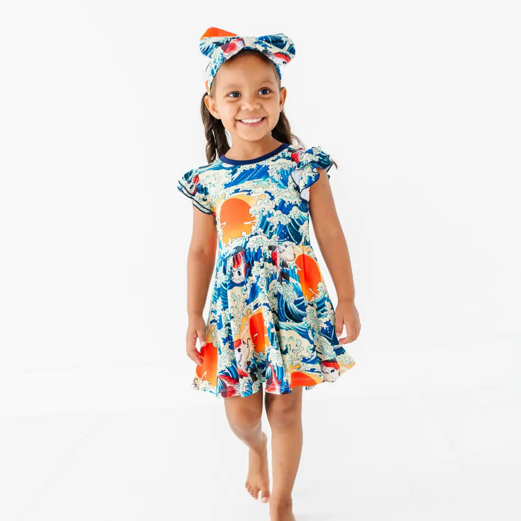 A smiling child walking in a flutter sleeve twirl dress and matching bow headband with a koi fish, wave, and sunset print.