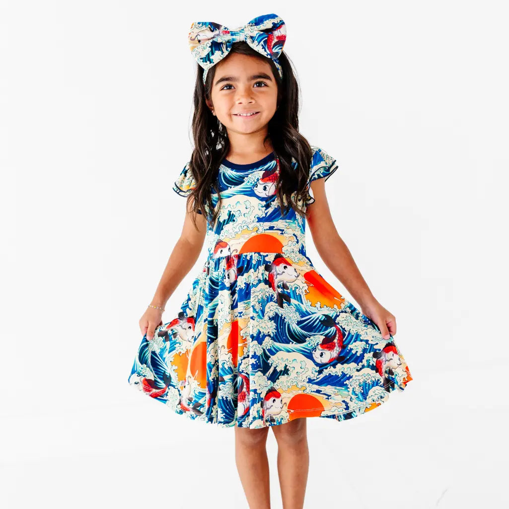 A child in a matching bow headband holding out both sides of a twirl dress printed with koi fish, ocean waves, and suns.