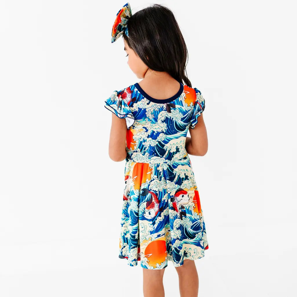 Back view of a child in a flutter sleeve twirl dress and bow headband with a koi fish, ocean wave, and sunset print.