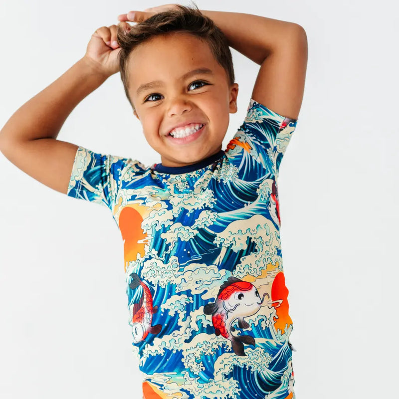 A smiling child with arms raised behind the head wearing a short sleeve pajama top with a koi fish, ocean wave, and sunset print.