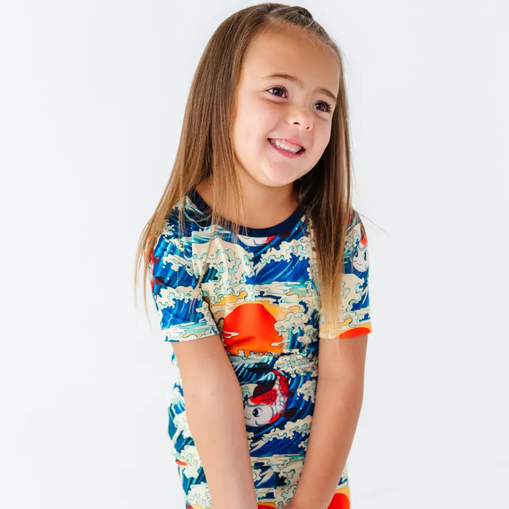 A smiling child glancing to the side wearing a short sleeve pajama top with a koi fish, ocean wave, and sunset print.