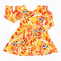 Flat lay of a long sleeve twirl dress with flutter ruffles at the shoulders in a golden yellow print of lion dance heads and red lanterns on a white background.