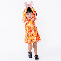 A toddler with pigtails smiling with both hands raised overhead, in a long sleeve twirl dress with flutter ruffles at the shoulders in a golden yellow print of lion dance heads and red lanterns.