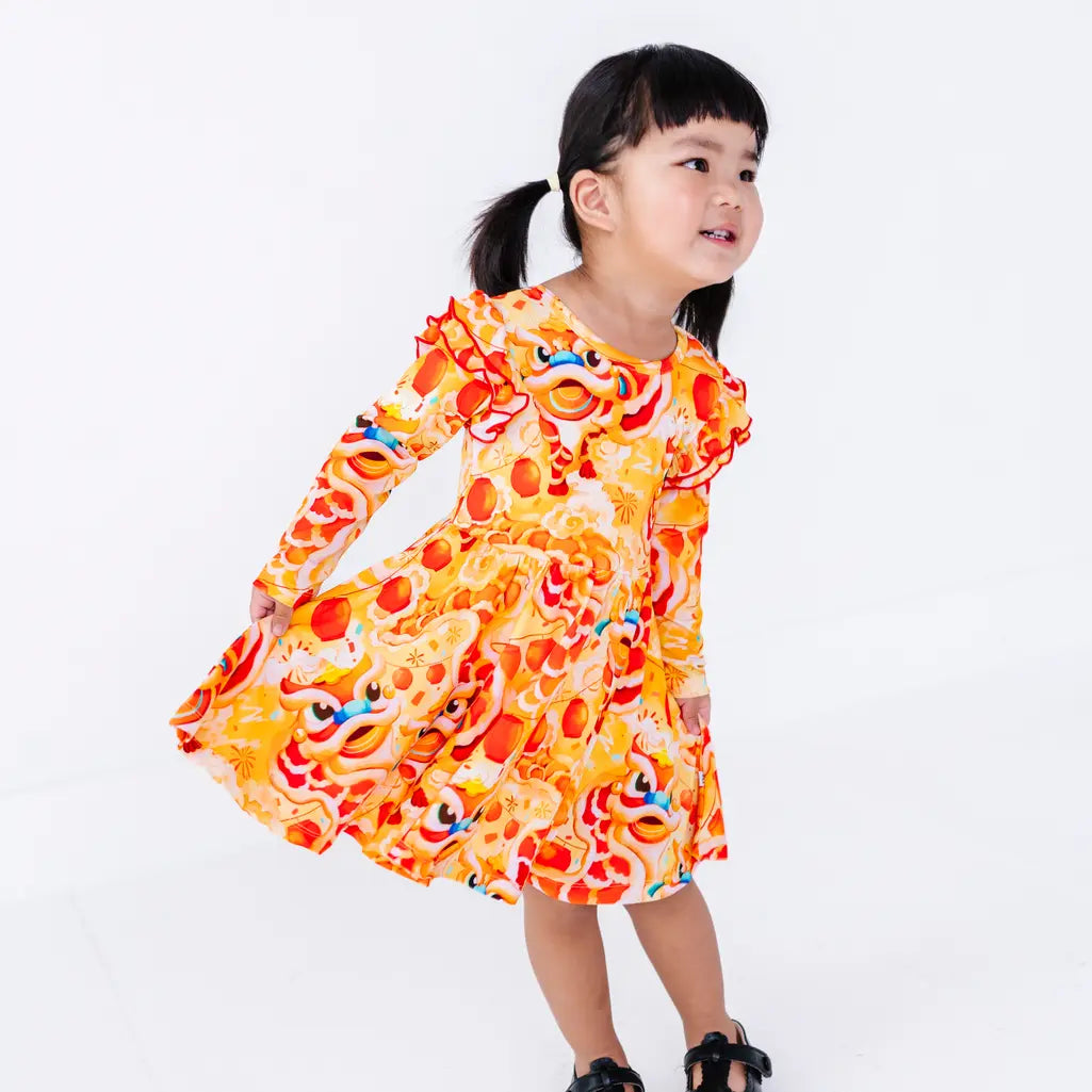 A toddler with pigtails looking aside and holding out the skirt of a long sleeve twirl dress with flutter ruffles at the shoulders, in a golden yellow print of lion dance heads and red lanterns.