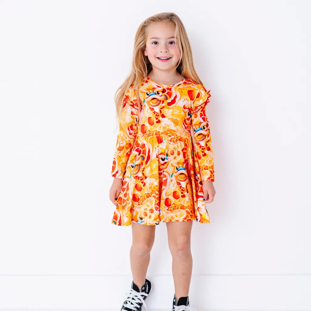 A child smiling at the camera and standing with arms down, in a long sleeve twirl dress with flutter ruffles at the shoulders in a golden yellow print of lion dance heads and red lanterns.