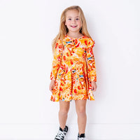 A child smiling at the camera and standing with arms down, in a long sleeve twirl dress with flutter ruffles at the shoulders in a golden yellow print of lion dance heads and red lanterns.